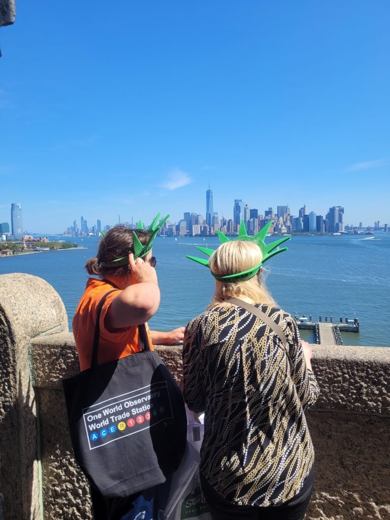 Two ladies wearing foam statue of liberty crowns on pedestal level viewing out into Hudson river with Manhattan in background. 